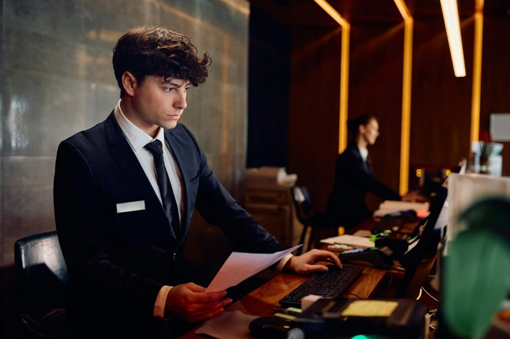 Male receptionist working on desktop PC at hotel front desk.