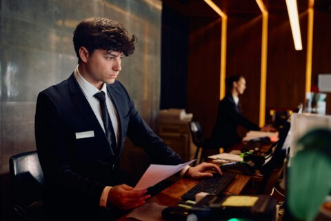 Male receptionist working on desktop PC at hotel front desk.