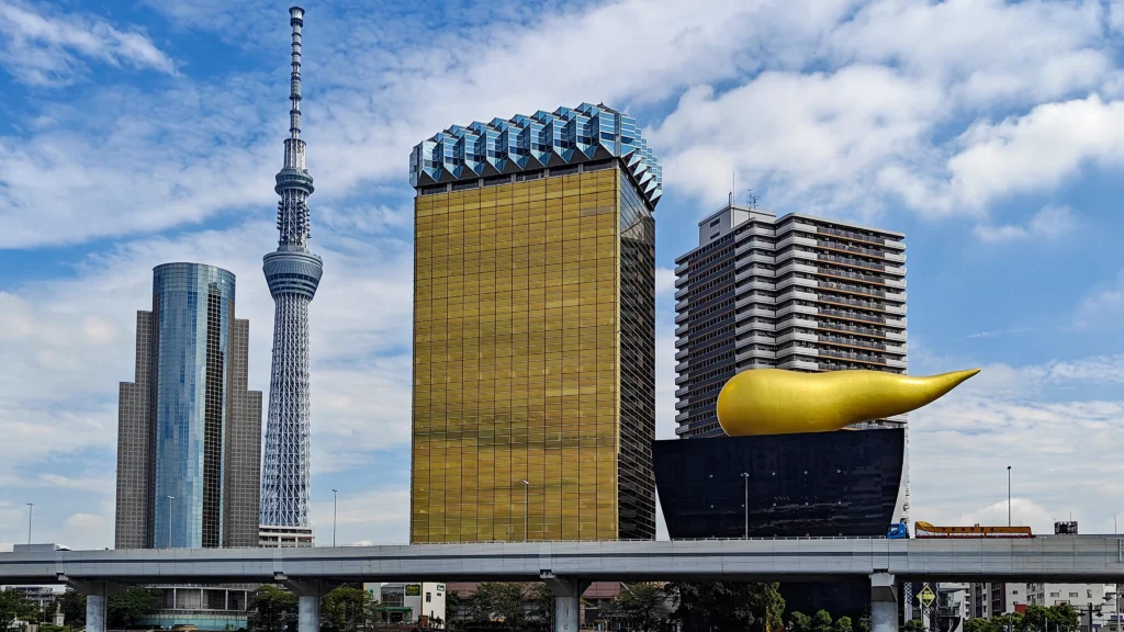 A cityscape showcasing the Tokyo Skytree, a symbol of Tokyo Triumph, stands tall beside modern buildings like the golden Asahi Beer Hall with its iconic flame sculpture under Japan’s vibrant blue sky.