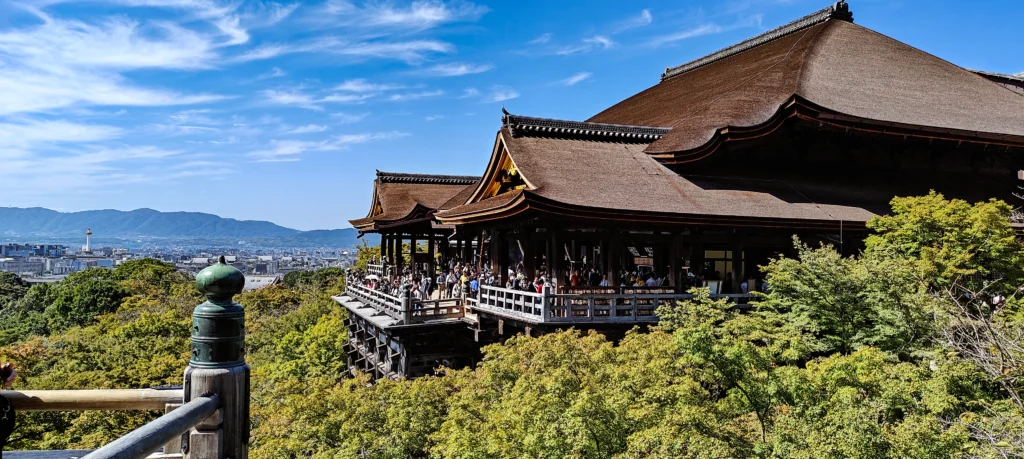 A large wooden temple with curved roofs, like those near Fushimi Inari, overlooks a green forest and city under a bright blue sky; many people stand on the balcony enjoying the view.