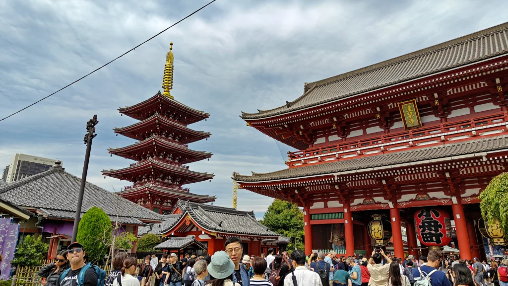 A crowd of people gathers in front of the historic Senso-ji Temple in Tokyo, Japan, with vibrant red buildings and a five-story pagoda under a partly cloudy sky—a true Tokyo Triumph.