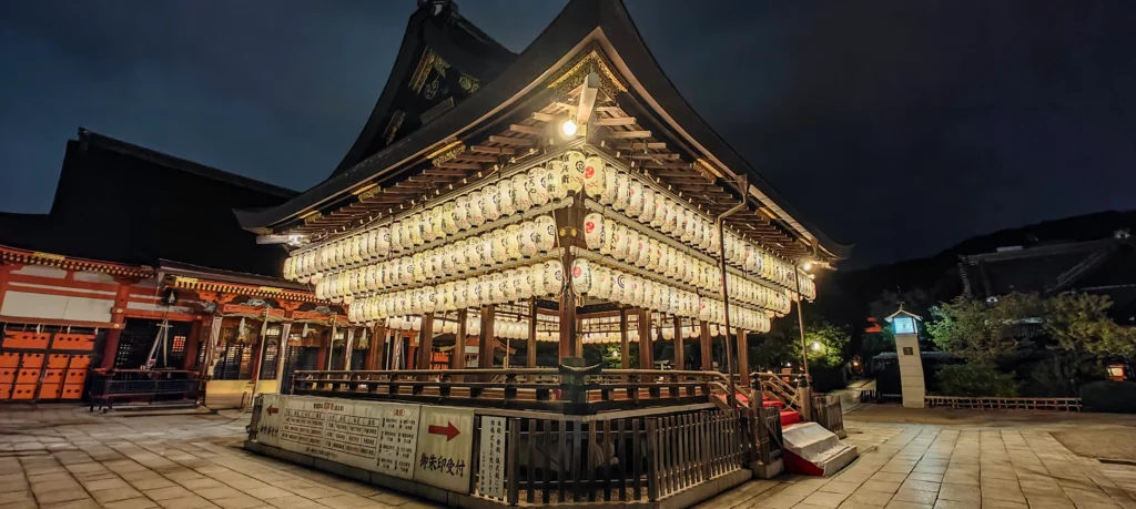 A traditional Japanese shrine at night in Kyoto, illuminated by rows of glowing paper lanterns hanging from its roof, with the dark sky and surrounding buildings of the Gion District in the background.