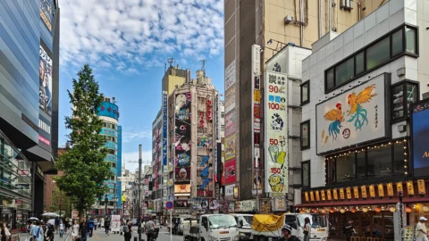 A busy city street in Tokyo, Japan, with colorful buildings covered in signs and advertisements, people walking, cars and trucks parked along the road—a perfect Solo Sojourn under a partly cloudy sky.